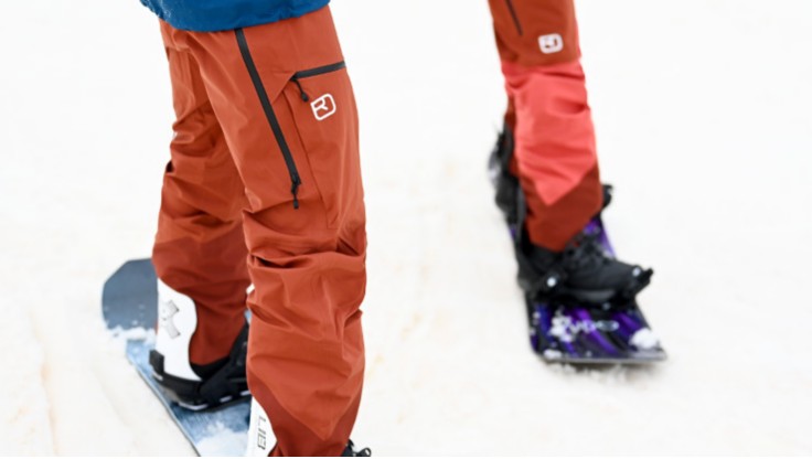 Un snowboarder debout dans la neige en pantalon à bretelles.