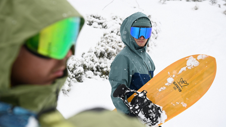 Ein Snowboarder steht am Gipfel und trägt eine Shell-Jacke.
